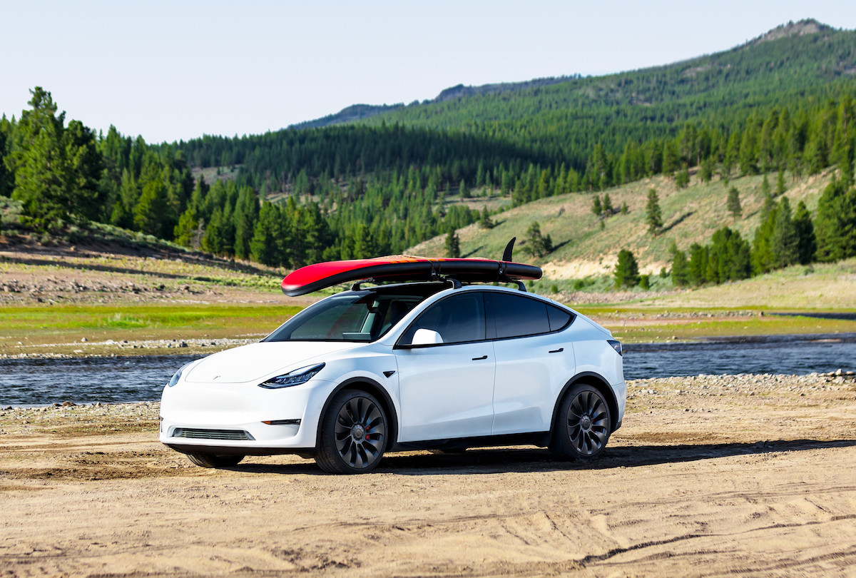 Tesla Model Y with a paddle board on the roof rack. White car, on a beach in mountain area